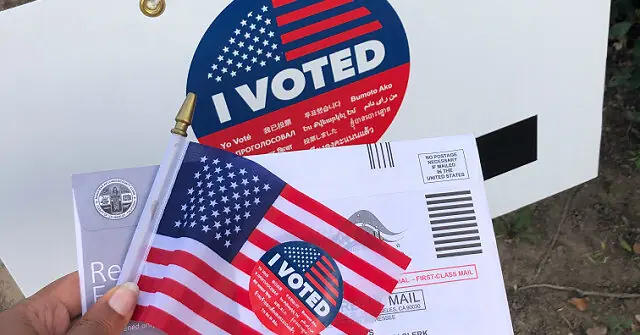 The image shows a hand holding a "I Voted" sticker and a mail-in ballot envelope with an American flag design, symbolizing the voting process in the United States.