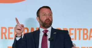 The image shows a man in a suit and tie speaking and gesturing with his right hand, with a backdrop featuring the word "Breitbart."