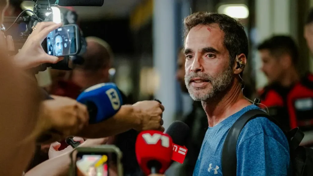 A man in a blue shirt is surrounded by reporters and microphones, appearing to be giving an interview or statement.