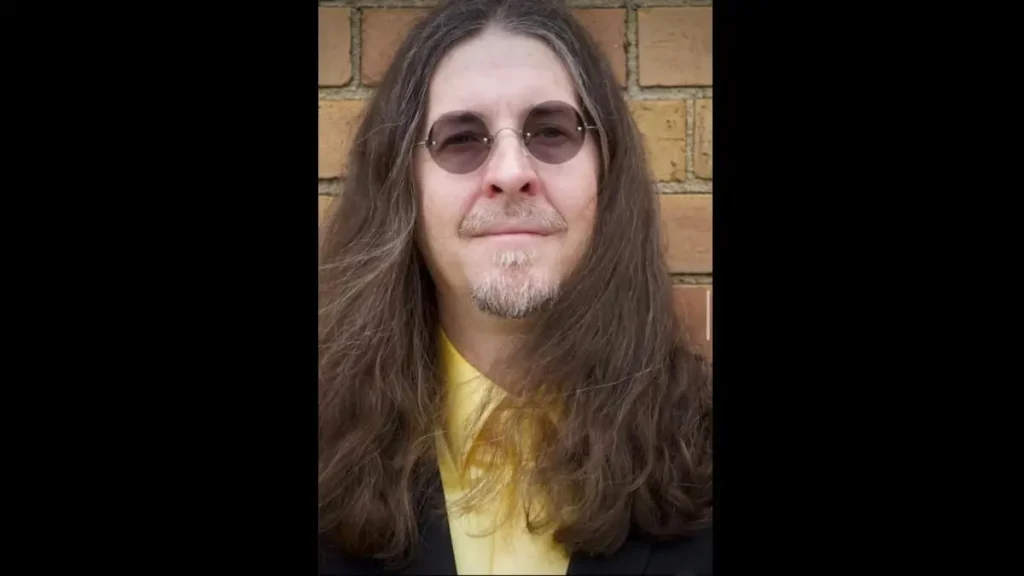 The image shows a person with long hair and glasses, wearing a yellow shirt and a dark jacket, standing in front of a brick wall.