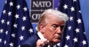 The image shows a person in a suit standing in front of a backdrop featuring the NATO logo and American flags.