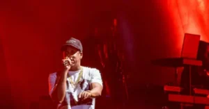 A performer is on stage, holding a microphone and wearing a cap and a chain, with red lighting illuminating the scene.