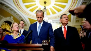 A group of individuals, including a man in a blue suit, are gathered in a formal setting, possibly a government or legislative building, with a chandelier and ornate ceiling in the background.