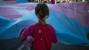 A young person carries a stuffed animal and a red backpack while participating in a protest or demonstration, holding a large trans flag with messages written on it.