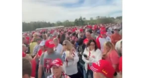 The photo shows a large crowd of people gathered outdoors, many wearing red hats and shirts, suggesting a political rally or public event.