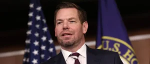 The image shows a man in a suit and tie speaking in front of an American flag and a U.S. House of Representatives flag.