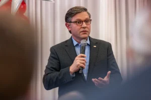 A man in a suit is speaking into a microphone, gesturing with his hand, in what appears to be a formal or professional setting.