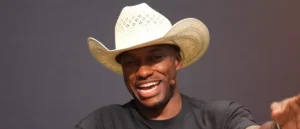 A man wearing a white cowboy hat smiles while speaking, with a microphone clipped to his shirt.