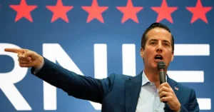 A man in a suit is speaking into a microphone while pointing to the side, with a backdrop featuring red stars and the letters "RNE."