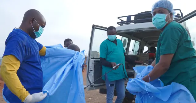 Medical personnel are seen preparing for a procedure or handling medical equipment outdoors near a vehicle.