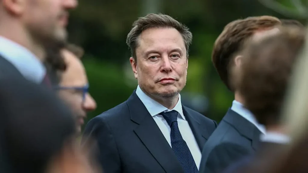 The image shows a man in a suit and tie standing outdoors, surrounded by other individuals.