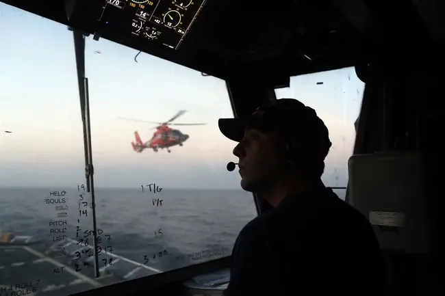 A person is seen inside a cockpit, observing a helicopter flying over the ocean.