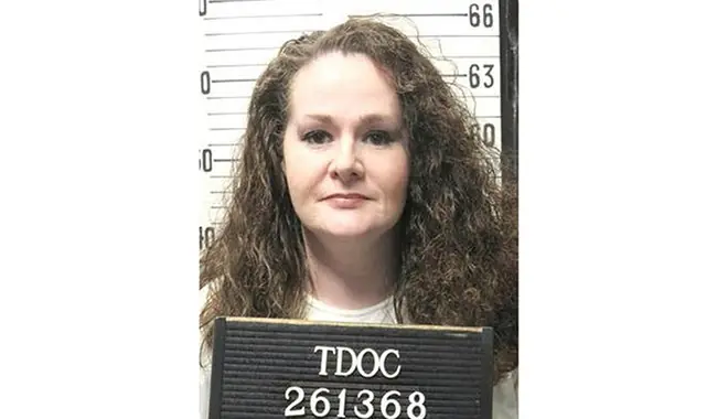 The image shows a mugshot of a person with long, curly hair, wearing a light-colored shirt, and holding a sign with "TDOC 261368" on it.