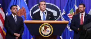 A man stands at a podium with the Department of Justice seal, delivering a statement in front of flags and other officials.