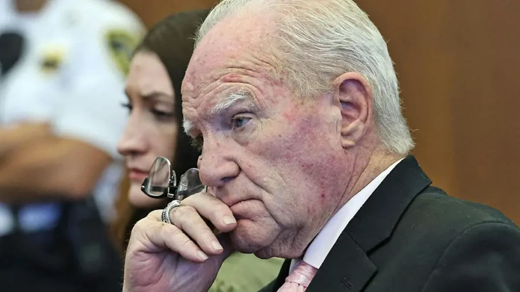 The image shows a man in a suit and tie, appearing contemplative, with a woman and a person in a white mask in the background.