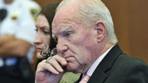 The image shows a man in a suit and tie, appearing contemplative, with a woman and a person in a white mask in the background.