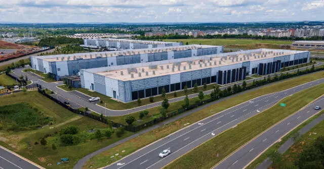 The image shows an expansive industrial complex surrounded by greenery and a highway, with a backdrop of a suburban area and distant hills under a partly cloudy sky.