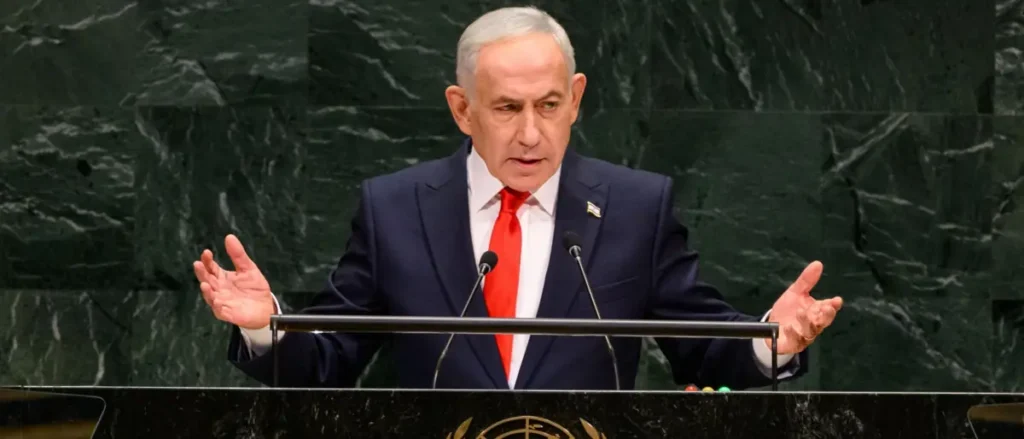 The image shows a man in a suit and red tie speaking at a podium with microphones, likely at a formal international event.