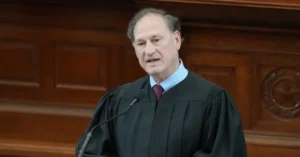 The image shows a person in a judicial robe standing at a podium, likely delivering a speech or making an announcement in a formal setting.