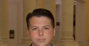 The image shows a person in a formal setting, possibly a government or institutional building, with columns and a door in the background.
