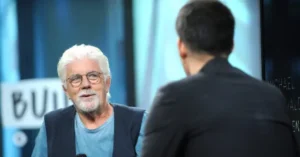 The image shows a man with white hair and a beard, wearing glasses and a blue shirt, engaged in a conversation with another person whose back is to the camera.