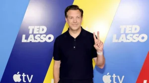 The image shows a person posing in front of a backdrop featuring the "Ted Lasso" logo and the Apple TV+ logo, making a peace sign with their hand.