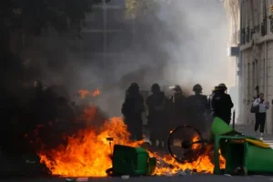 A group of police officers in riot gear stand near a burning barricade during a protest.