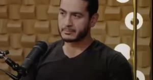 The image shows a person speaking into a microphone in a studio setting with acoustic foam panels in the background.