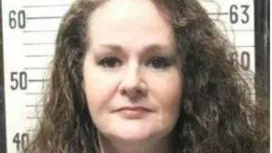 The image appears to be a mugshot of a person with long, curly hair, standing in front of a height measurement scale.