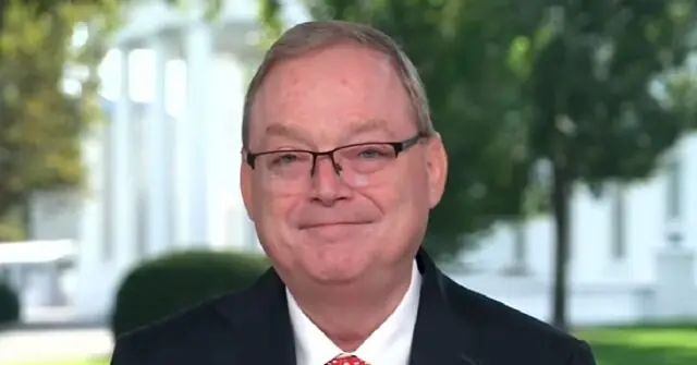 The image shows a man in a suit and tie, smiling, with a blurred background of greenery and white structures.