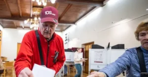 An elderly man wearing a red jacket and a cap is seen at a polling station, holding a piece of paper, while another person in a blue jacket is assisting him.