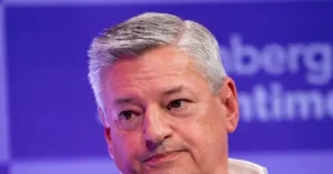 The image shows a close-up of a person with gray hair, set against a blue background with partially visible text.