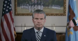 The image depicts a man in a suit and tie, seated in front of an American flag and a framed painting, suggesting a formal or official setting.