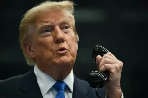 The image shows a person in a suit holding a microphone, appearing to be speaking or addressing an audience.