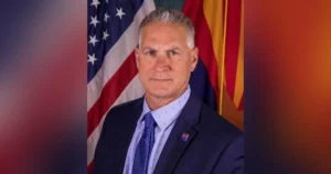 The image shows a man in a suit and tie standing in front of the American flag and another flag, likely representing a political or official setting.