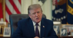 The image shows a person in a formal suit seated at a desk, with an American flag and other flags in the background, suggesting a formal or official setting.