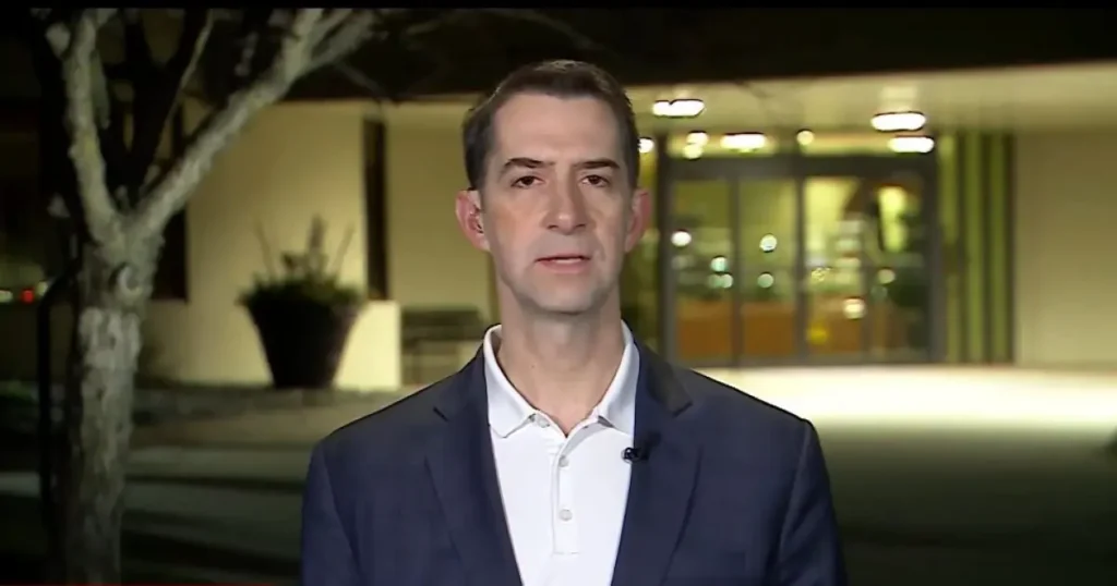 The image shows a man in a suit standing in front of a building at night, with a tree and some indoor lighting visible in the background.