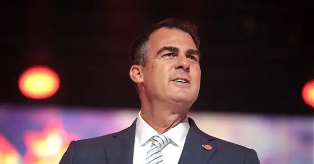 The image shows a man in a suit and tie, speaking at what appears to be a formal event or conference, with a colorful background.