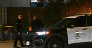 Two police officers are seen near a police vehicle at a crime scene, with crime scene tape and a fence in the background.