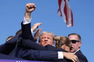 The image shows a political figure raising his fist in a gesture of defiance or solidarity, with an American flag visible in the background.