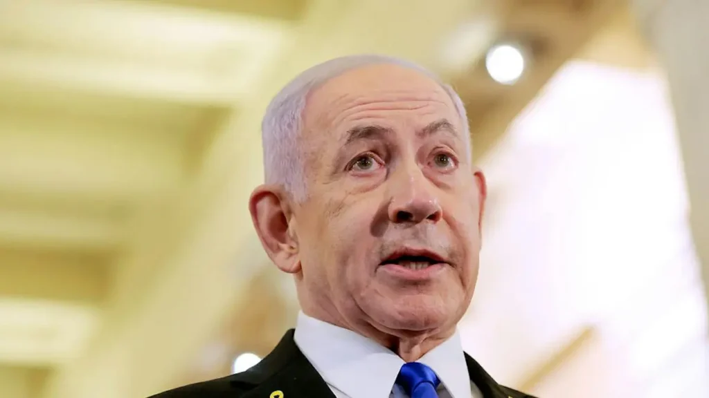 The image shows a man in formal attire, including a suit and tie, speaking in what appears to be a professional or political setting.