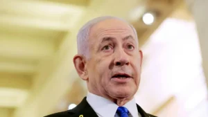 The image shows a man in formal attire, including a suit and tie, speaking in what appears to be a professional or political setting.