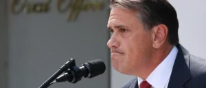 A man in a suit and tie is speaking into a microphone at an outdoor event.