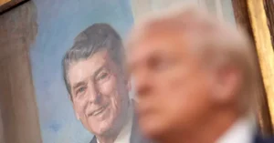 The image shows a close-up of a person's face in the foreground, partially obscuring a portrait of a smiling man in the background.