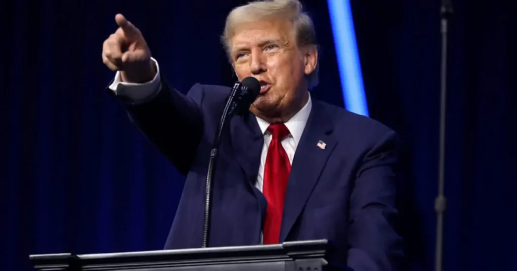 The image shows a person in a suit and tie speaking at a podium, with one arm raised and pointing.