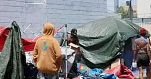 The image shows a group of individuals in a makeshift outdoor living area with tents and belongings, suggesting a homeless or refugee camp setting.