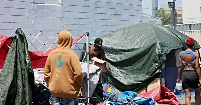 The image shows a group of individuals in a makeshift outdoor living area with tents and belongings, suggesting a homeless or refugee camp setting.