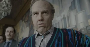 The image depicts a man in a colorful striped sweater and a bow tie, standing in a room with a chandelier and a mural in the background.