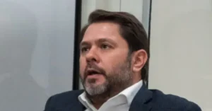 The image shows a man with a beard and short hair, wearing a suit and a white shirt, speaking in what appears to be a professional or formal setting.