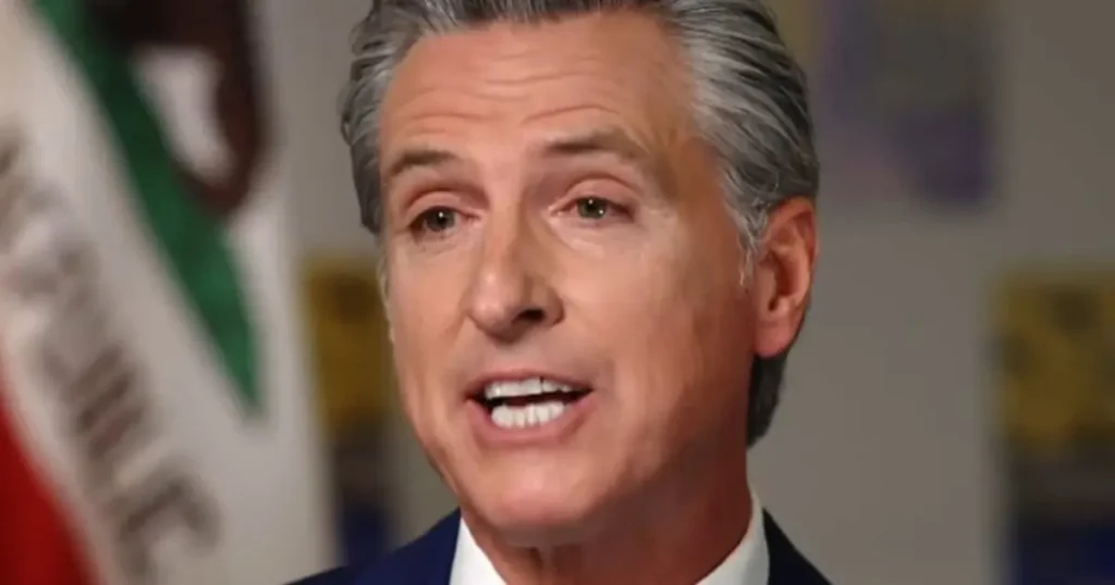 The image shows a man in a suit speaking in front of a blurred background that includes a flag.